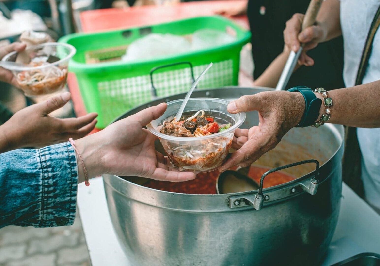 Two people serving food from a large pot into clear plastic bowls at an outdoor setting.
